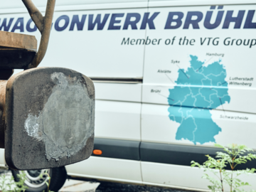 Eine Nahaufnahme eines Güterwaggons, im Hintergrund ein weißer Transporter mit der Aufschrift 'Waggonwerk Brühl, Member of the VTG Group'. Auf dem Transporter ist eine Deutschlandkarte abgebildet, die verschiedene Standorte der VTG-Waggonwerke zeigt.